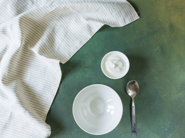 On a green background, there is an empty round bowl in the center of the photo. Next to it is a spoon, a bowl of sour cream and a napkin. You can put any dish on the plate. Horizontally. Top view, flat layout - Photo, image