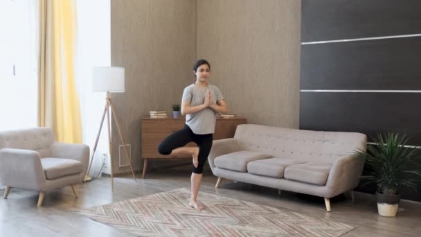Fiatal indiai nő meditál áll a szőnyegen és a kézen Namaste, csinál egyensúly gyakorlat jóga, sportruházat Light Room Otthon reggel - Felvétel, videó