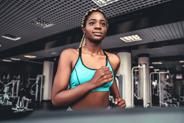 Молодая улыбающаяся африканка занимается фитнесом и тренировками во время бега по треку в спортивном клубе. Потеря веса и здоровье в тренажерном зале - Фото, изображение