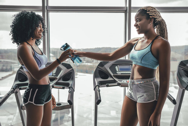 Generous girl giving bottle of water to the friend in gym. Sporty lifestyle. Side view of two black women on training - Fotó, kép