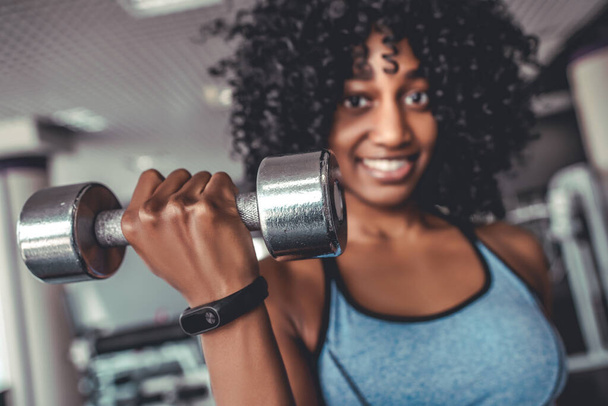 Portrait of smiling african women looking camera in gym lifting dumbbell. Yong black sportive girl with curly hair on training biceps - Valokuva, kuva
