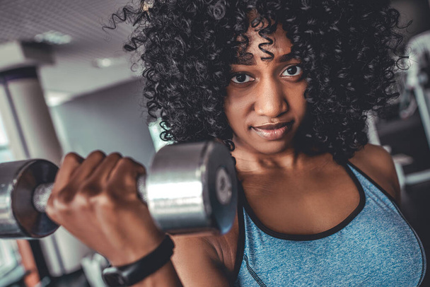Portrait of happy healthy young american woman afro hairstyle working out in a gym with a pair of dumbbells - Zdjęcie, obraz