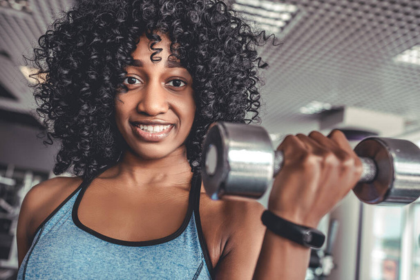 Portrait of cheerful african young woman with activity tracker on hand doing exercise with dumbbels in gym. Muscular body and wellbeing concept - Фото, изображение