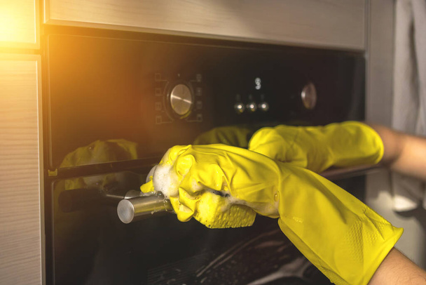 Wycieranie i czyszczenie drzwi piekarnika w kuchni domowej z żółtą gumową rękawicą i gąbką - Zdjęcie, obraz