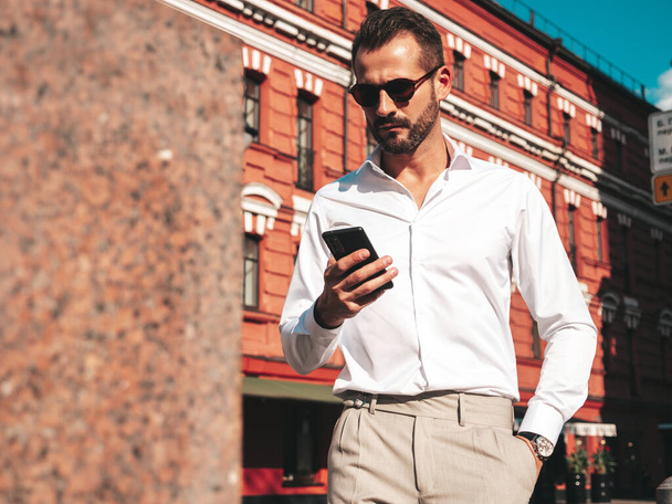 Handsome confident stylish hipster lambersexual model.Modern man dressed in white shirt and trousers. Fashion male posing in the street in Europe city at sunset. In sunglasses. Looking at smartphone - Foto, imagen