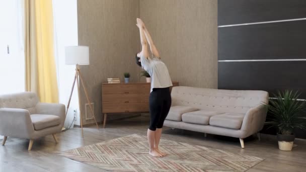 Genç bir kadın yoga yapıyor, vücudunu esnetme egzersizi yapıyor, sırtını geriyor, spor kıyafetleri giyiyor, evdeki parlak bir odada. - Video, Çekim