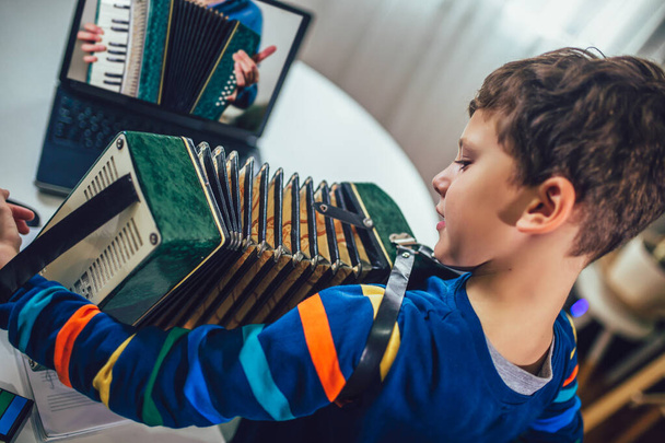 Keskittynyt poika pelaa harmonikkaa ja katselee verkkokurssia läppärissä harjoitellessaan kotona. Verkkokoulutus, verkkokurssit. - Valokuva, kuva