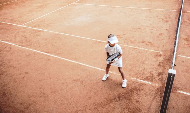 Ребёнок играет в теннис на открытом глиняном корте. Вид сверху маленького теннисиста на открытый теннисный корт. Полный снимок спортивной девочки на теннисных тренировках в клубе. Активные занятия для детей. - Фото, изображение