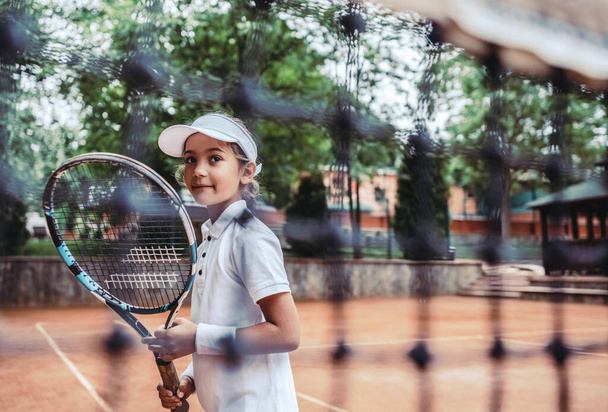 Little girl with tennis racket in sport club. Cropped image of a sporty little girl with racket playing tennis outdoors. Child athlete on tennis court. Training for young kid. - 写真・画像