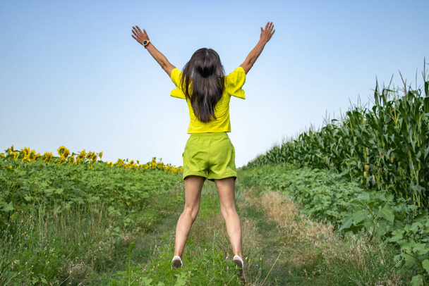 Glückliche junge Frau in gelber Bluse mit langen Haaren breitet ihre Hände aus. Sie springt in einem Feld aus Sonnenblumen und Mais gegen den blauen Himmel. Es geht um Spiritualität und Freiheit. Konzept Spiritualität und Freiheit. - Foto, Bild