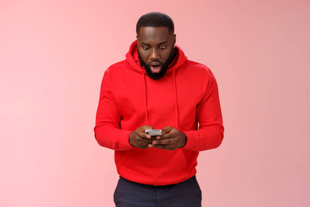 Amused focused impressed african-american bearded guy in red hoodie drop jaw widen eyes look amazed smartphone display winning hard game level answering exciting message, standing pink background - Photo, Image