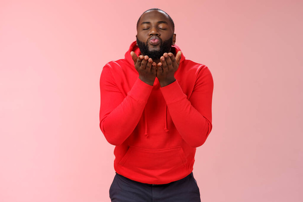 Χαριτωμένο χαριτωμένο αφροαμερικανός γενειοφόρος τύπος σε κόκκινο hoodie κλειστά μάτια πτυσσόμενα χείλη κρατήστε τις παλάμες κοντά στο στόμα στέλνοντας παθιασμένο φιλί αέρα φυσώντας mwah κάμερα, στέκεται υπέροχο ροζ φόντο - Φωτογραφία, εικόνα