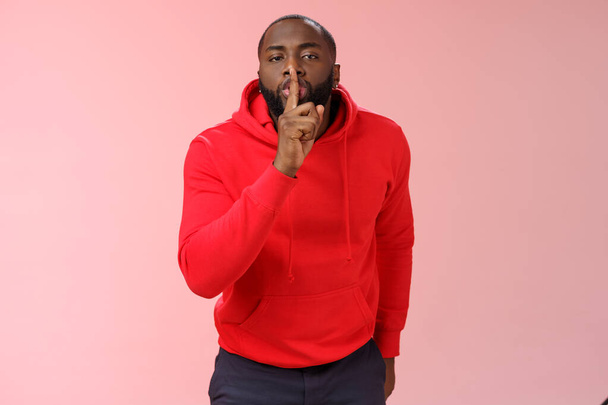 Shhh it taboo. Portrait sensual funny cute african-american bearded man in red hoodie bending camera show shush gesture index finger on lips telling secret asking keep quiet not tell anyone mystery - Photo, Image