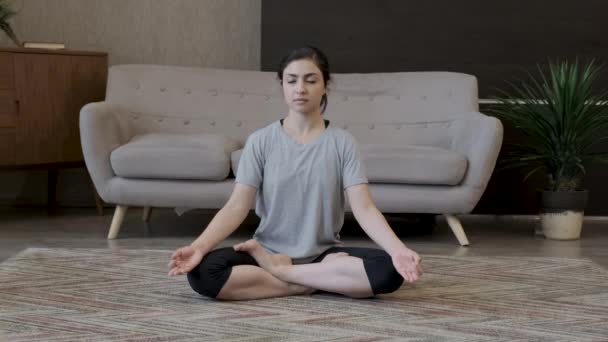Eine junge Inderin sitzt in einer Yoga-Lotus-Pose, ein Mädchen macht eine Übung, sitzt auf dem Boden, trägt einen Hut und ein graues T-Shirt und ist in einem gemütlichen Raum zu Hause - Filmmaterial, Video
