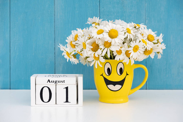 August 1st date on calendar and bouquet of chamomile in yellow mug with funny face against blue wall. Hello August summer month concept. - Foto, afbeelding