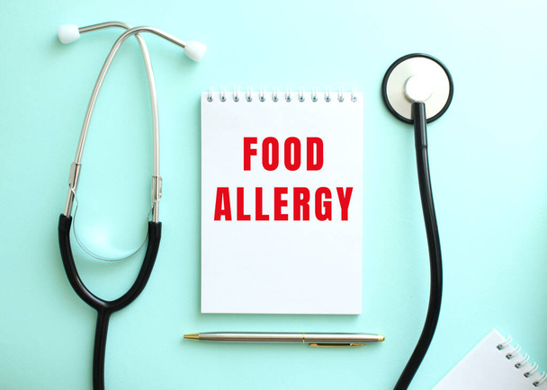 On a blue background, a stethoscope and a white notepad with the red words FOOD ALLERGY. Medical concept - Φωτογραφία, εικόνα