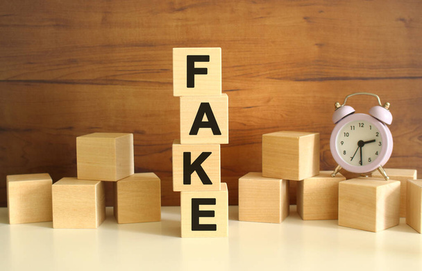 Four wooden cubes stacked vertically on a brown background form the word FAKE. Cubes are scattered nearby and there is a clock. Front View Concepts - Foto, Bild