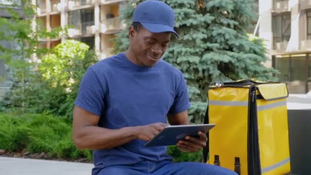 Sorrindo afro-americano homem mensageiro sentado no banco usa tablet olhando câmera - Filmagem, Vídeo