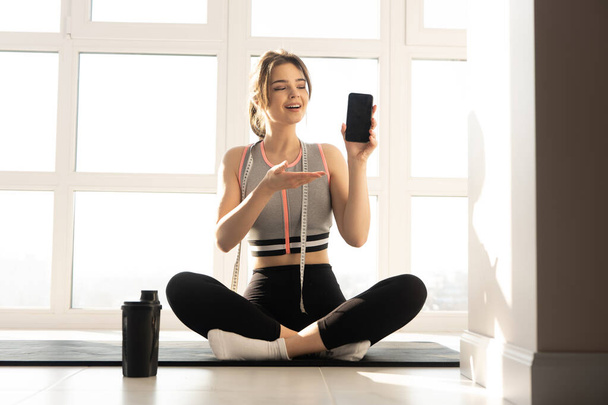 Sportovkyně ukazující mobilní telefon na fitness podložce - Fotografie, Obrázek