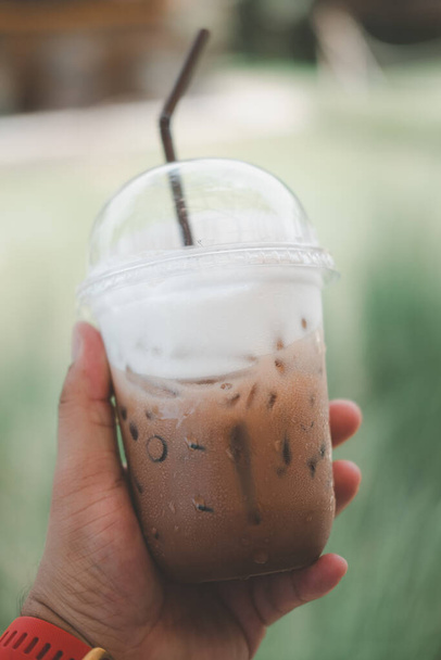 Закройте руки молодой азиат держа в руках пластиковую чашку вкусного кофе мокко со льдом на красивом размытом зеленом фоне природы. - Фото, изображение