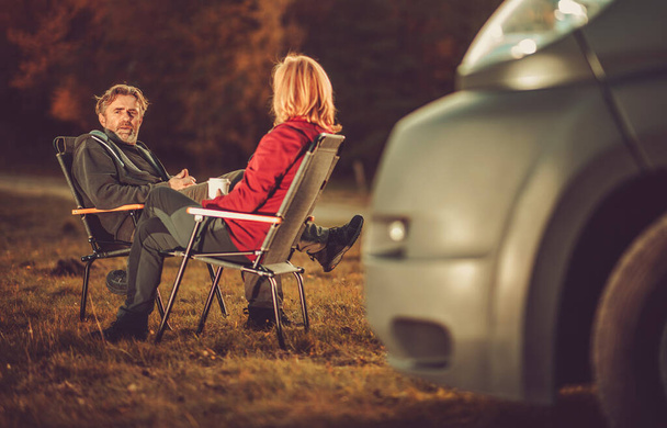 Hombres y mujeres caucásicos charlando en un camping junto a Camper Van. Asientos en sillas de campamento en algún lugar remoto. - Foto, Imagen