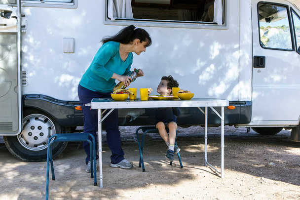 Η μαμά σερβίρει φαγητό στο γιο της μπροστά από το τροχόσπιτο, στο τραπέζι στη σκιά του δέντρου.. - Φωτογραφία, εικόνα
