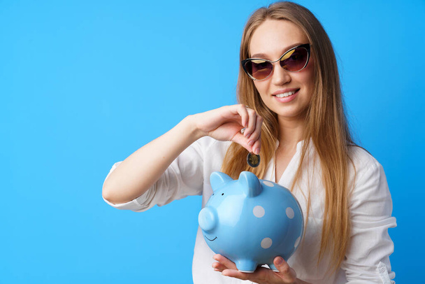 Beautiful smiling young woman with piggy bank on blue background - Foto, Bild