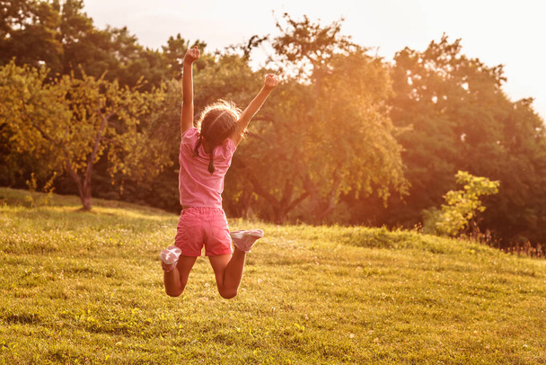 Pikkutyttö hyppää korkealle kädet ylhäällä. Lapsi iloitsee kauniina kesäpäivänä. Onnellinen lapsi kesäiltana - Valokuva, kuva