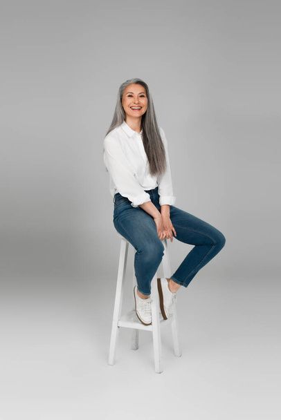 cheerful asian woman in blue jeans and white shirt sitting on high stool on grey background - Zdjęcie, obraz