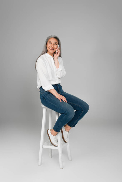 cheerful asian woman in jeans sitting on high stool and talking on smartphone on grey background - 写真・画像