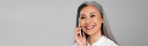 mujer asiática feliz con el pelo gris llamando en el teléfono móvil aislado en gris, bandera - Foto, imagen