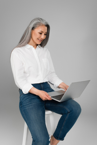 happy asian woman in white shirt typing on laptop while sitting on stool isolated on grey - Valokuva, kuva