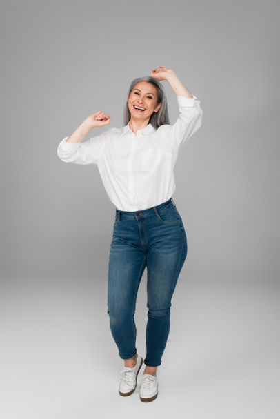 joyful asian woman in jeans and white shirt showing yeah gesture on grey background - Valokuva, kuva