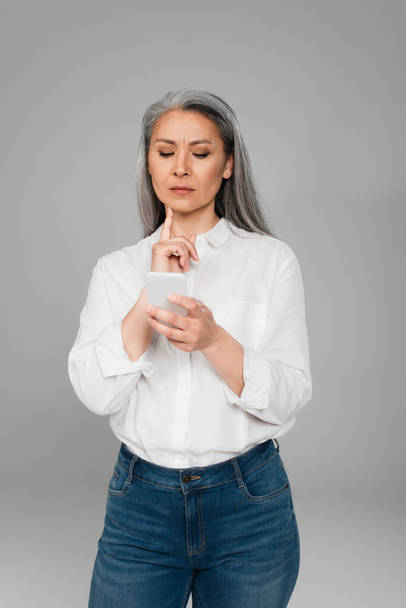 thoughtful asian woman touching chin while looking at cellphone isolated on grey - Фото, зображення
