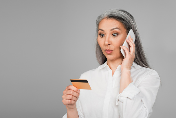 zaskoczona azjatycka kobieta dzwoni na telefon komórkowy patrząc na kartę kredytową odizolowany na szary - Zdjęcie, obraz