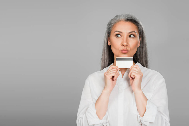 thoughtful asian woman in white shirt holding credit card isolated on grey - 写真・画像