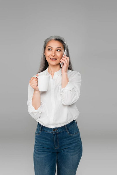 positive asian woman holding cup of tea while talking on mobile phone isolated on grey - Foto, Imagen