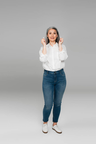 full length view of mature asian woman in jeans and shirt holding earphones on grey - Zdjęcie, obraz