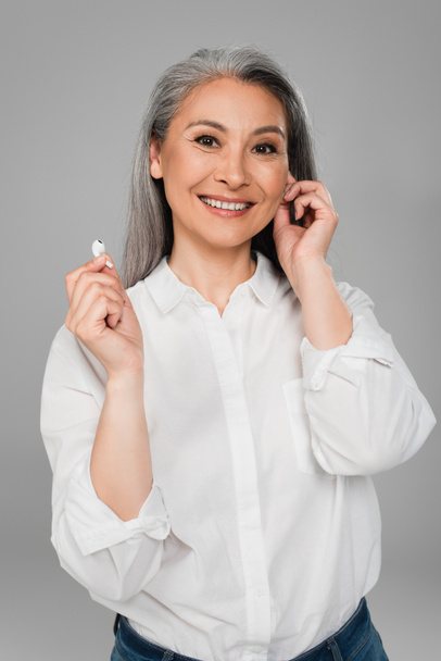 boldog ázsiai nő fehér ing gazdaság vezeték nélküli fülhallgató elszigetelt szürke - Fotó, kép