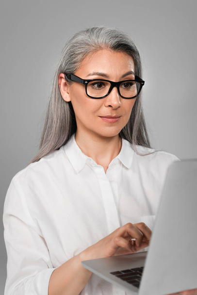 volwassen aziatische vrouw in wit shirt en stijlvolle bril typen op laptop geïsoleerd op grijs - Foto, afbeelding