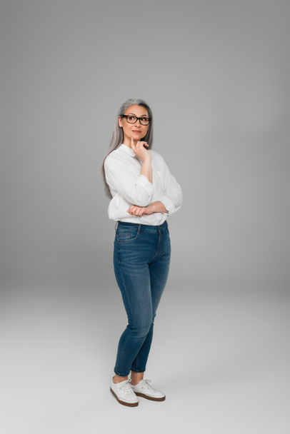 full length view of mature asian woman in jeans, white shirt and eyeglasses standing on grey  - Foto, immagini