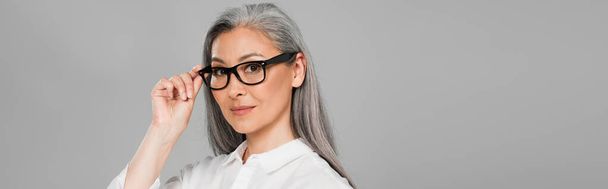 mature asian woman with grey hair adjusting eyeglasses isolated on grey, banner - Foto, imagen