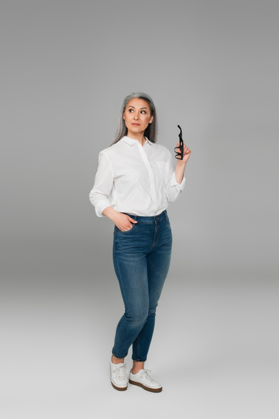 middle aged asian woman holding eyeglasses while standing with hand in pocket of jeans on grey  - Foto, Imagem