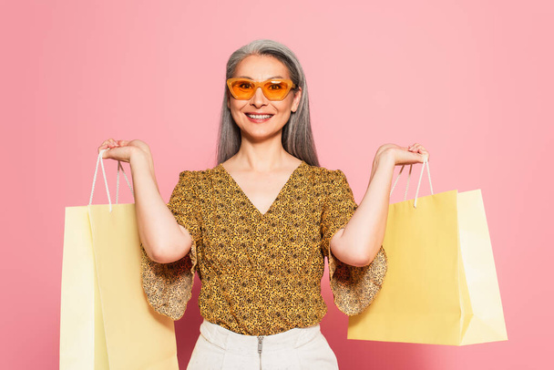 cheerful asian woman in stylish clothes holding purchases on pink background - 写真・画像