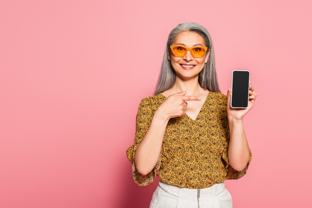 smiling asian woman in stylish sunglasses pointing at smartphone on pink background - Fotografie, Obrázek