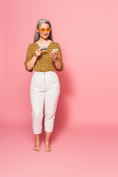 full length view of trendy asian woman with credit card and cellphone on pink  - Photo, Image