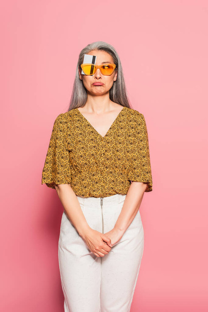 mature asian woman in yellow sunglasses, with eye covered with credit card grimacing on pink  - Φωτογραφία, εικόνα