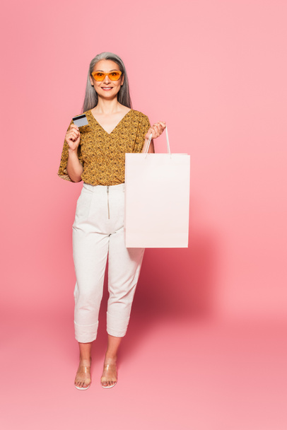 stylish asian woman in white pants standing with credit card and shopping bag on pink background - Foto, Imagen