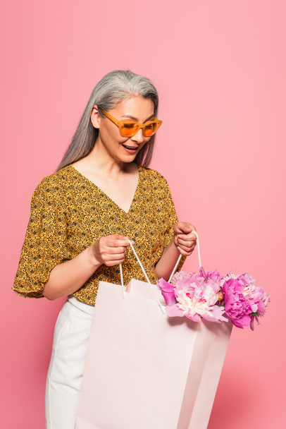 amazed asian woman looking into shopping bag with peonies isolated on pink - Fotoğraf, Görsel