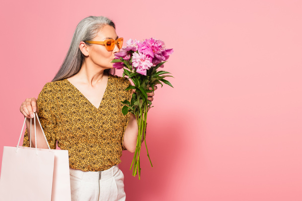 mature asian woman with shopping bag enjoying aroma of fresh peonies on pink background - 写真・画像